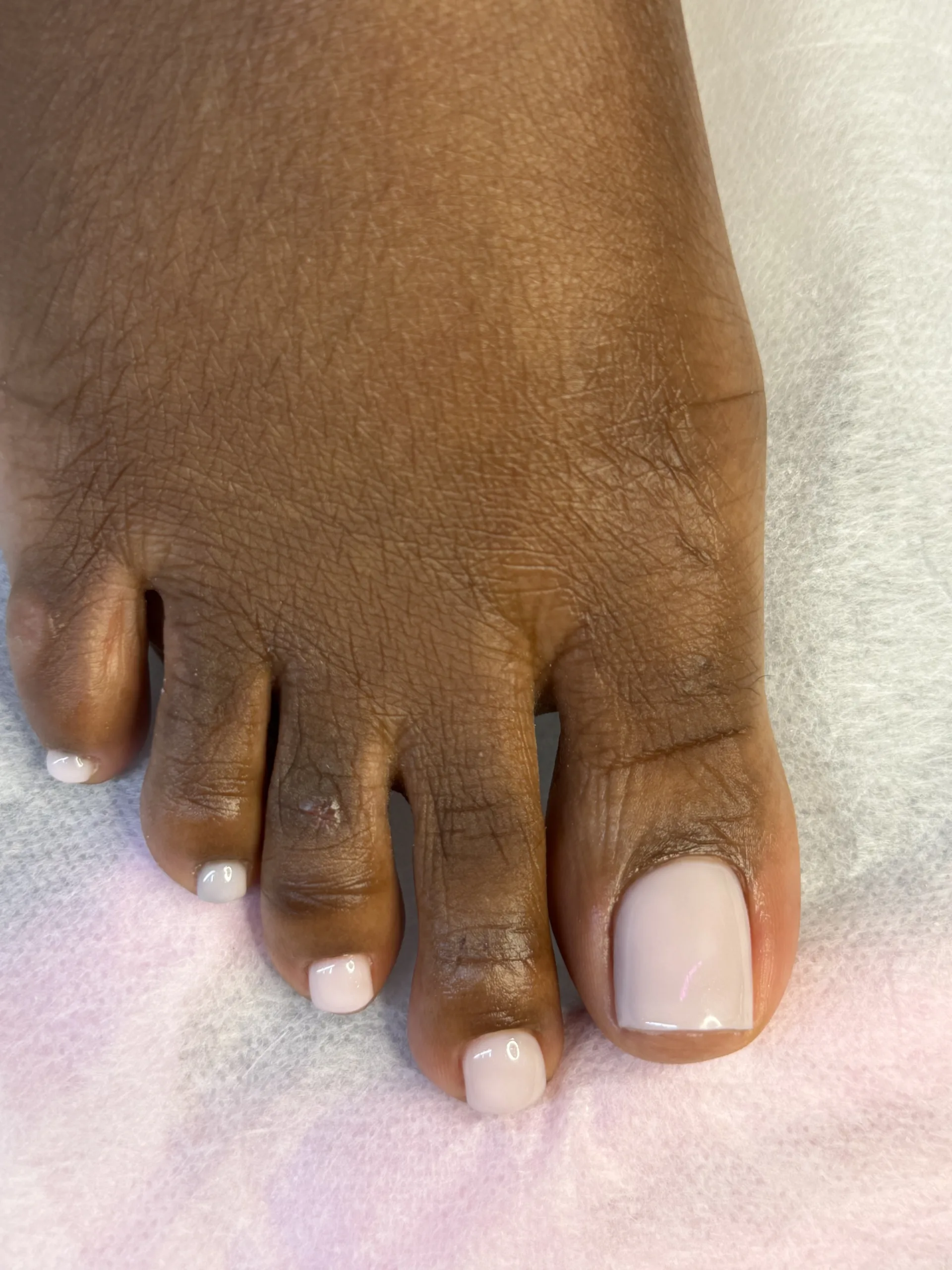 Soft milky nude pink pedicure on short toenails, shown in a close-up against deep skin tone